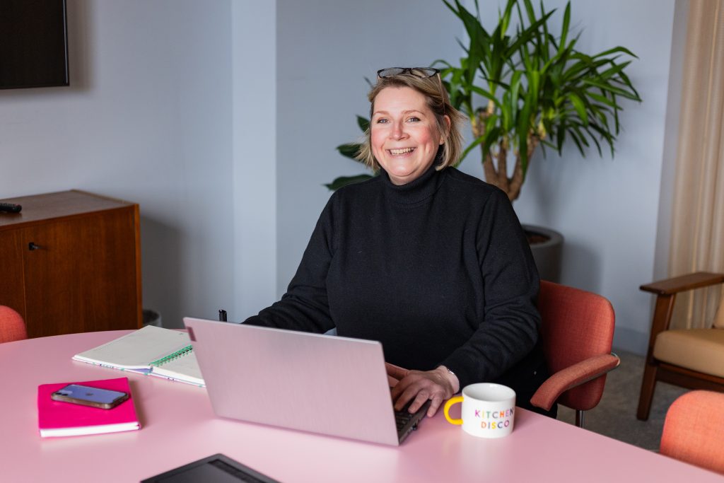 Jacqui Kemp looking up from her laptop screen, with a notebook, pen, smartphone, and a coffee mug on desk